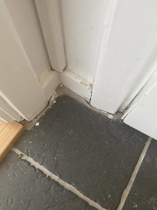 Skadad vit dörrkarm och foder möter en mörk stengolvplatta med en synlig söm och sprickor.