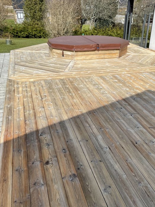Nybyggd träterrass med inbyggd rund jacuzzi täckt med brunt lock, i en solig trädgård.