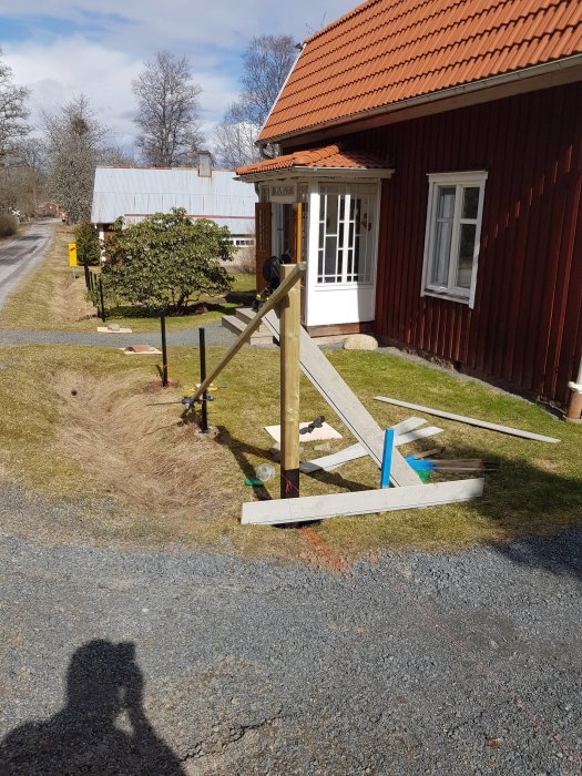 Nyligen uppsatta grindstolpar bredvid en röd stuga med verktyg och material på gräsmattan.
