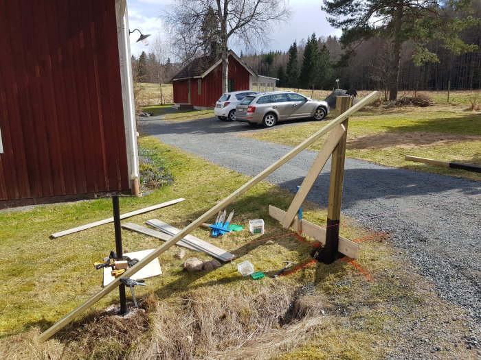 Två nyinstallerade trägrindstolpar på en gård, verktyg och material synliga på marken i förberedelse för vidare konstruktion.