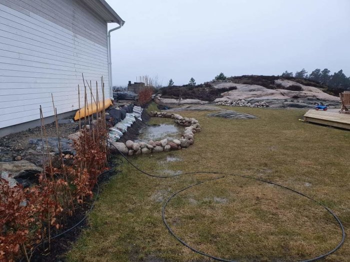 Blöt gräsmatta med pöl bildad efter regn, diket med stenar, slangen på marken vid husvägg.