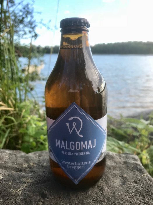 Flaska med etikett "MALGOMAJ Klassisk Pilsner IIA" från Västerbottens bryggeri, utomhus med sjö i bakgrunden.