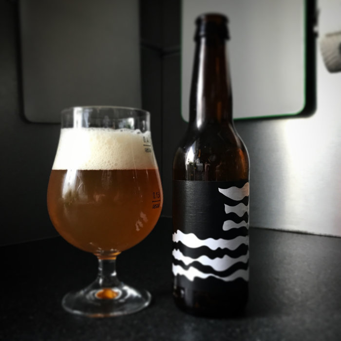 Ölglas fyllt med skummande öl bredvid en flaska med etikett, Omnipollo Nebuchadnezzar, på en köksbänk.