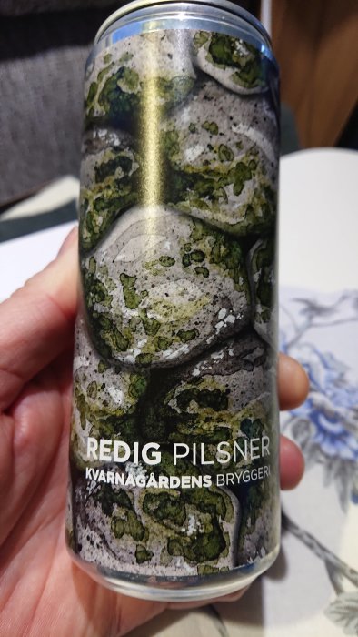 Hand håller en burk Redig Pilsner från Kvarnågardens Bryggeri med stenmönster.