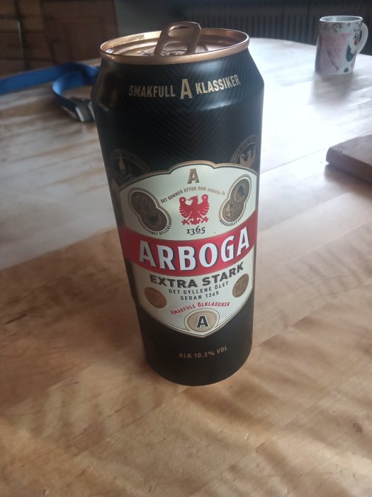 En öppnad burk av Arboga Extra Stark öl på ett träbord med en kopp i bakgrunden.
