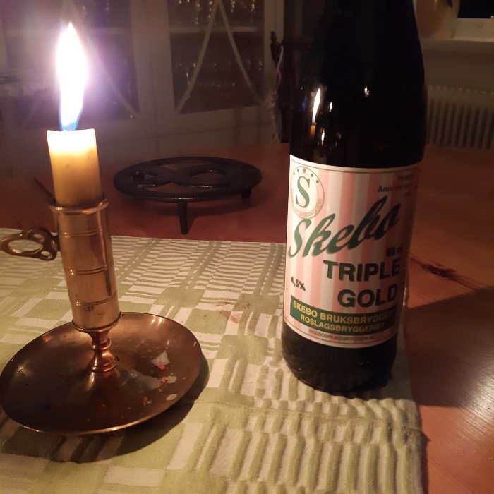 Flaska Skebo Triple Gold öl på bord med tänd ljus, i hemmiljö.
