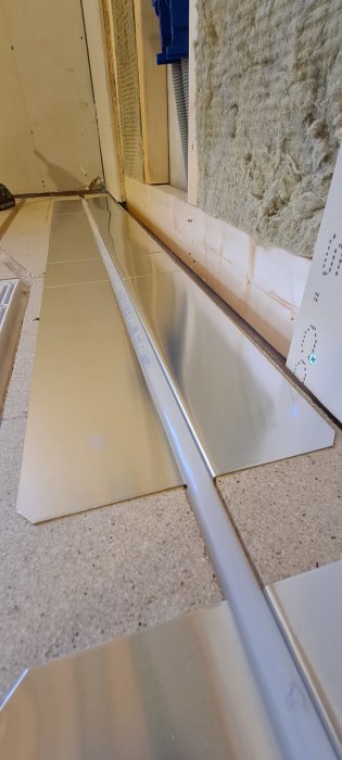 Metallplåtar installerade på ett golv med synliga luftfickor och monteringsskruvar, nära isoleringsmaterial.