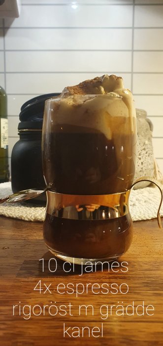 Ett glas med espresso och tjockt skum toppat med kanel på en träyta, med textrecept framför.