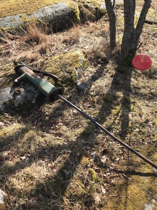 Gammal handdriven jordborr ligger på mossbeklädd mark med träd och stenar i bakgrunden.