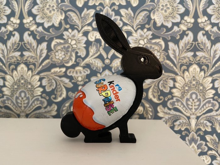 3D-utskriven kaninfigur håller ett Kinder Surprise-ägg mot ett blommigt tapetbakgrund.
