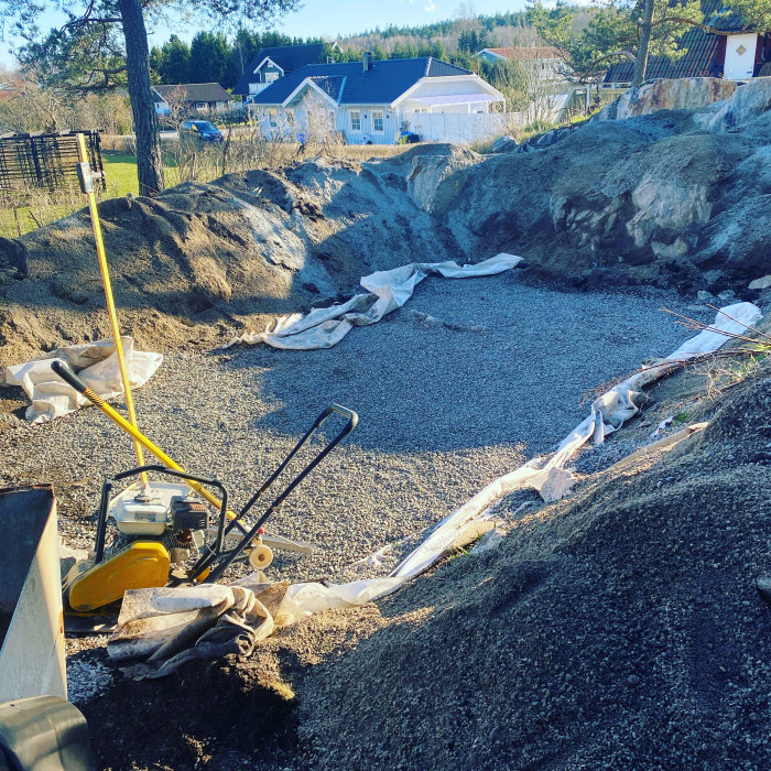 Gruslagd utgrävning för pool med jämnad botten och byggmaskin, omringad av berg och hus.