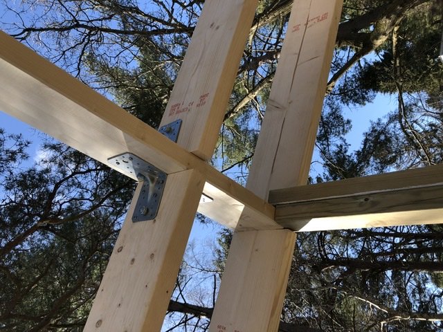 Trähörnkonstruktion med förstärkningsbeslag mot en bakgrund av träd och blå himmel.