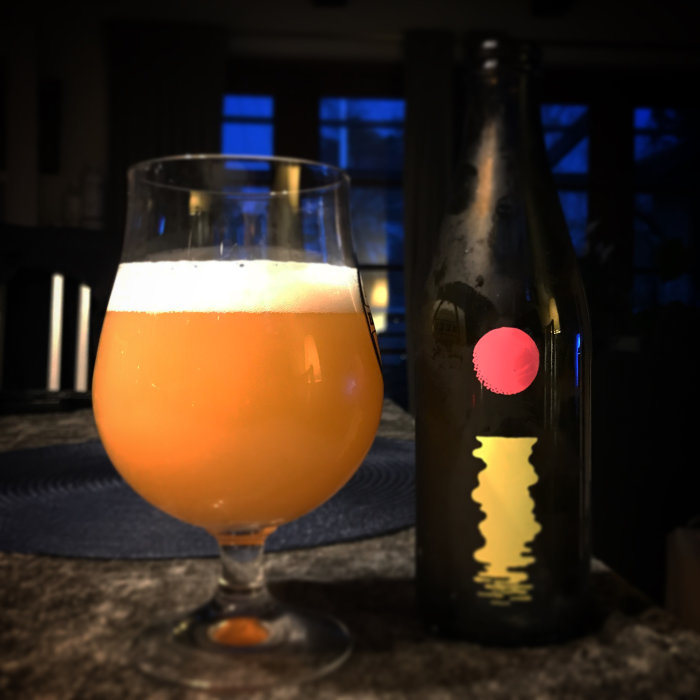 Ett glas med ölet Omnipollo Fatamorgana bredvid en tom flaska mot en suddig bakgrund.