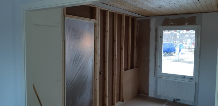 Delvis avtäckt vägg i renoveringsprojekt med synliga träreglar och bjälkar, bredvid ett fönster med utsikt utomhus.