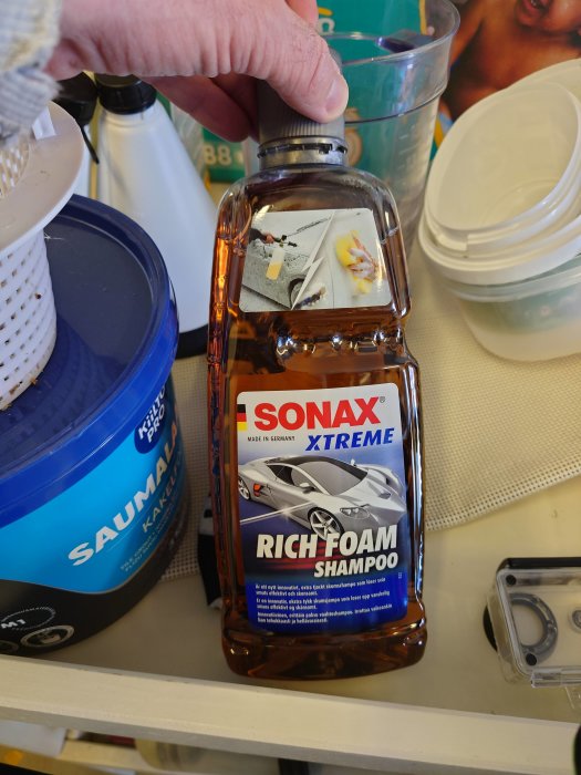 En hand håller en flaska Sonax Xtreme Rich Foam Shampoo för bilvård på en hylla med städprodukter.