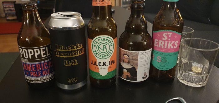 Fem olika ölflaskor och en burk, inklusive Poppel American Pale Ale, Black is Beautiful IPA, J.A.C.K. IPA, samt två andra öl, bredvid två tomma glas.