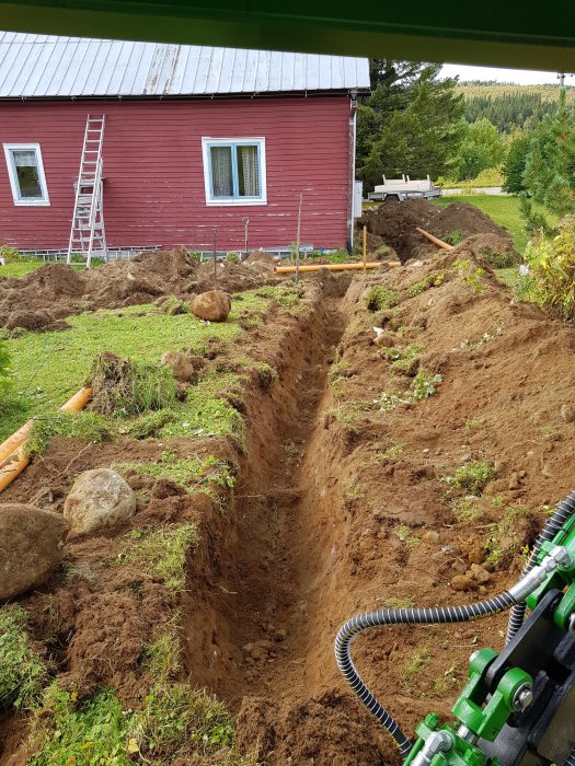 Grävd gröngrävning i trädgård med synliga rörledningar, röd stuga och stege i bakgrunden.