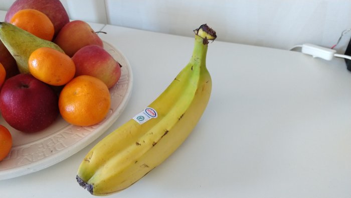 En ovanlig tvillingbanan ligger på ett bord framför en fruktskål med olika frukter.