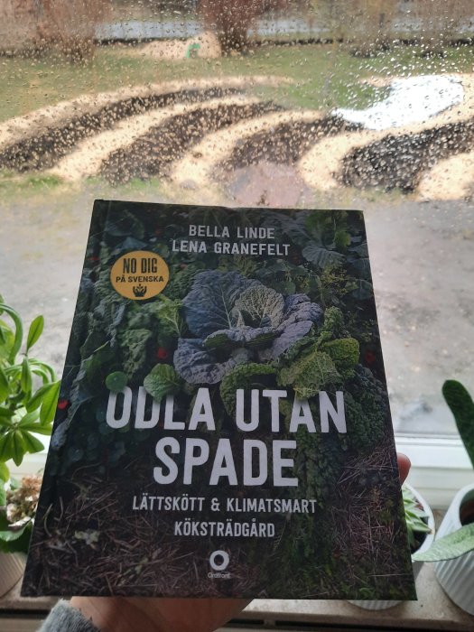 Hand håller boken "Odla utan spade" framför ett fönster med regndroppar och synlig trädgård i bakgrunden.