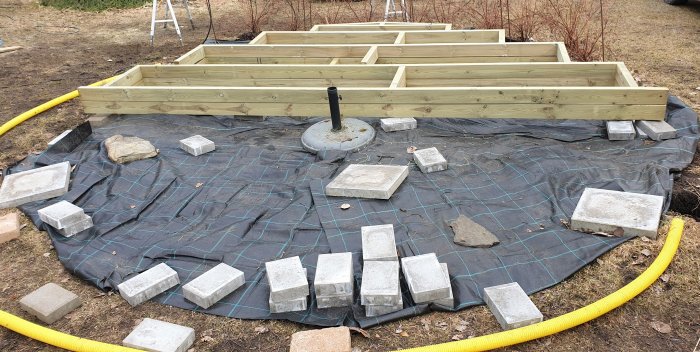 Konstruktion av en halvrund trädäcksram på markduk med stenplattor runt om och en gul slang.