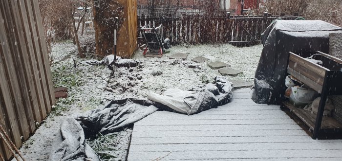 En påbörjad grävning i en trädgård täckt av snö, med trädgårdsredskap och vattenslang.