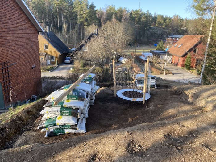 Trädgårdsprojekt med lerig jordhög, säckar av jord och torv, och ett trädomgärdat av skyddsnät.