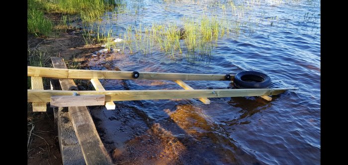 Brygga byggd av trä och däck, placerad vid en strandkant med vass och klart vatten.