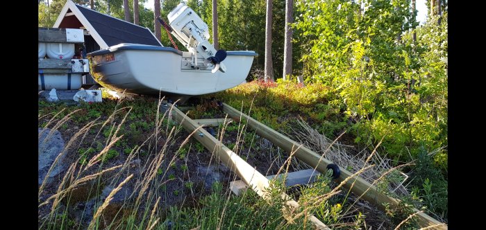 Båt med motor upphöjd på hemmagjord träkonstruktion med hjul i trädgård.