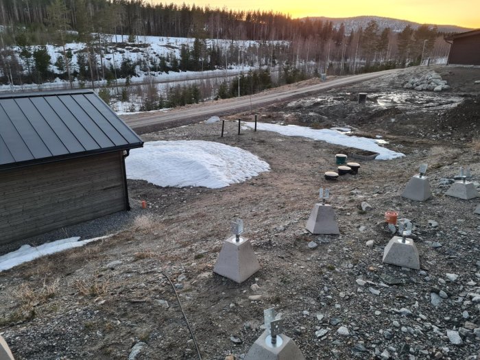 Nybyggt hus med grusad platt yta intill garage, övergång till sluttande tomt i skogsmiljö under arbete.