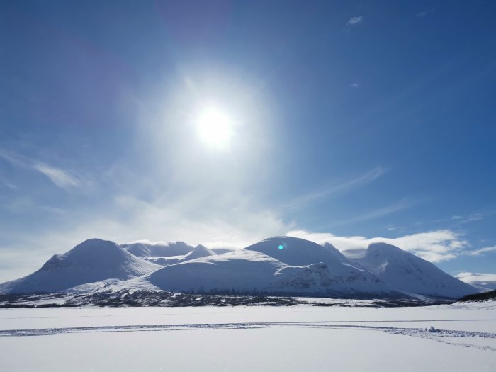 Solig vinterdag vid en fjällsjö med snötäckta berg och klarblå himmel.