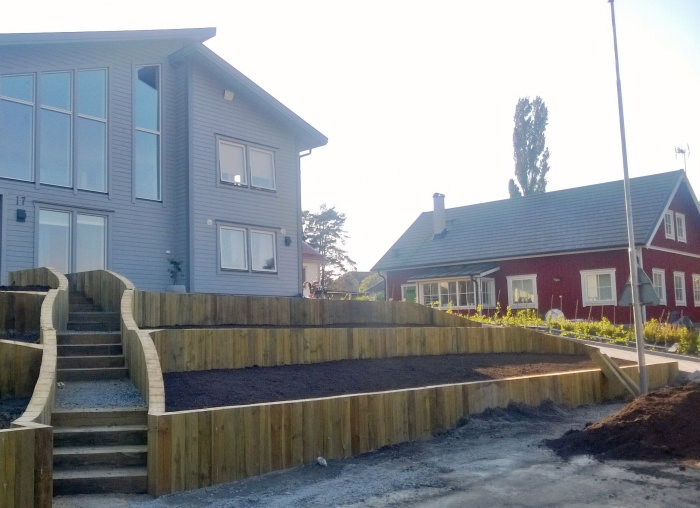 Grå husfasad med trästödmur i slänten framför, nyplanterade ytor och en trätrappa som leder uppåt.