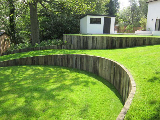 En välunderhållen trädgård med en krökt stödmur av byggtimmer som skapar terrasserad gräsmatta.