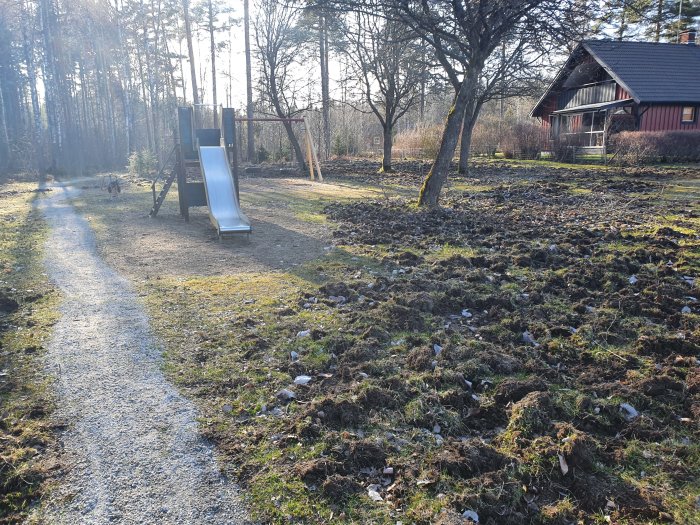 Trädgård med uppriven gräs och jord i förgrunden, lekstuga och trottoar i bakgrunden.