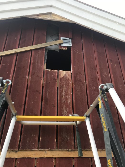 Stegen framför en husvägg med röd panel där ett hål till limträbalk är påbörjat, med synlig vindpapp bakom panelen.