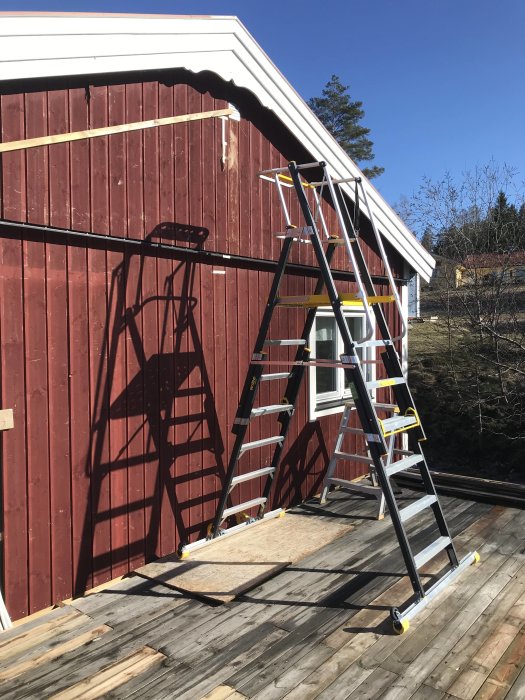 Röd träpanelvägg på hus med en stege uppställd och brädor på marken, klart för renovering.