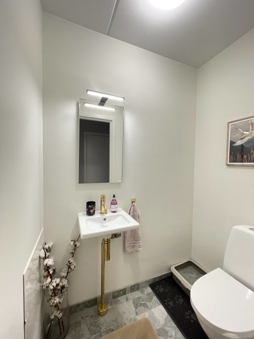 Litet badrum med vit inredning, guldig kran, toalett, spegel och en taklampa.