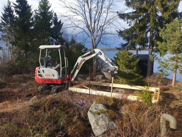 Minigrävare nära en trägjutform på byggplats med skogsområde och sjö i bakgrunden.
