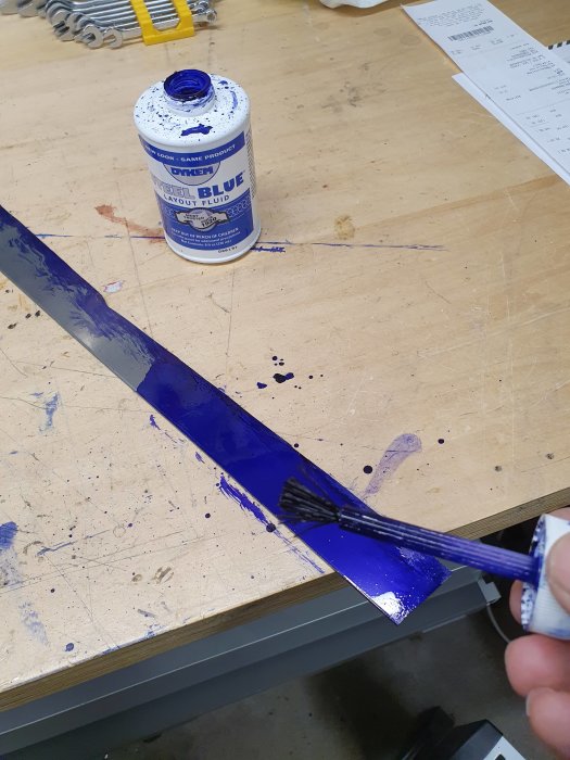 Stålremsa täckt med ritsvätska bredvid en öppen flaska och pensel som används för påstrykning på ett arbetsbord.