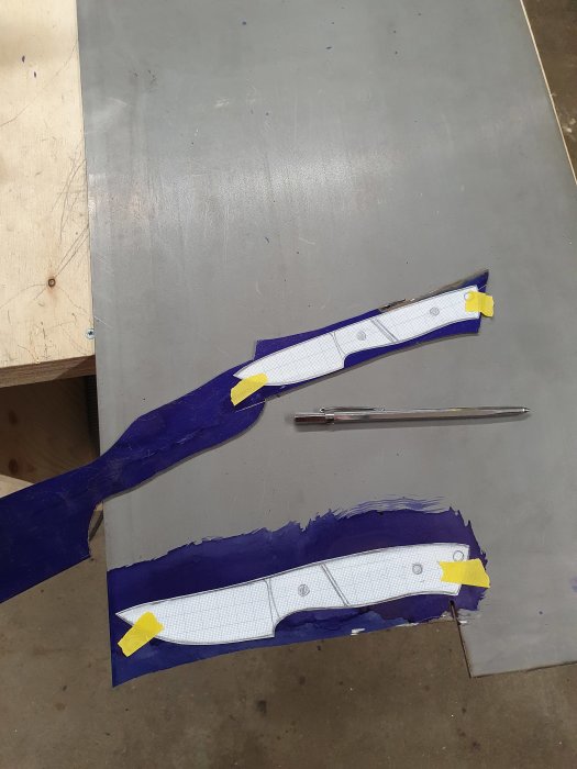 Två stålblad med påtejpade knivschabloner ligger på en metallplåt bredvid en ritpenna.