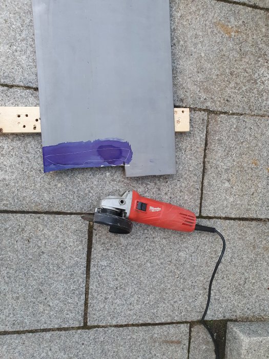 Vinkelslip bredvid en stor metallplåt med uppritad knivsform på stenläggning.