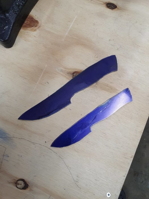 Två oråade knivblad med mörk ytbeläggning på en träbänk.