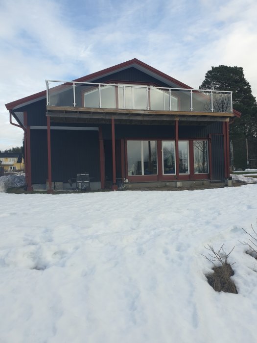 Röd trävilla med stor balkong på övervåningen, omgiven av snötäckt mark.