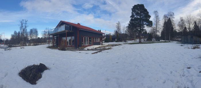 Panoramavy av rödmålat hus med balkong och stor öppen snötäckt trädgård.