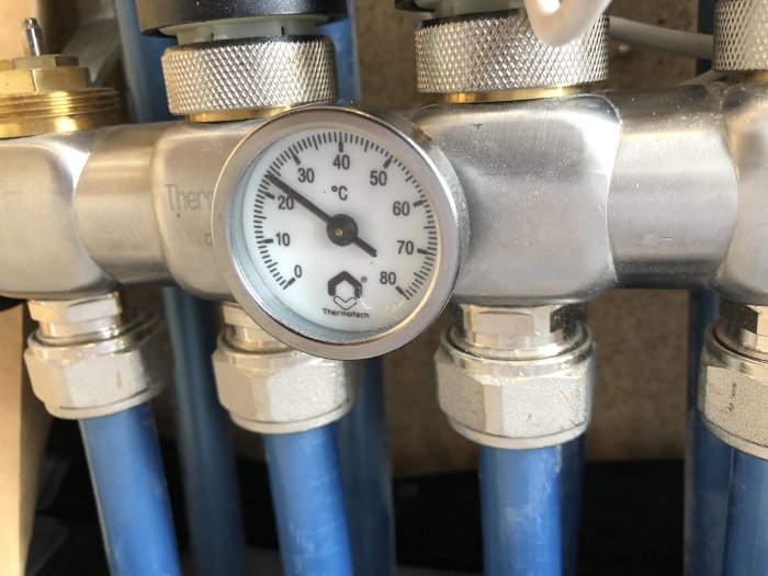 Termostat med vridbar ratt på golvvärmesystem visande temperatur runt 20 grader Celsius, monterad på rörledning.