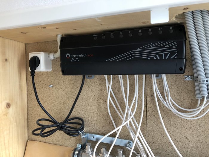 Uppkopplad Thermotech EC8-central för golvvärmesystem med synliga ledningar och rör under ett hyllplan.