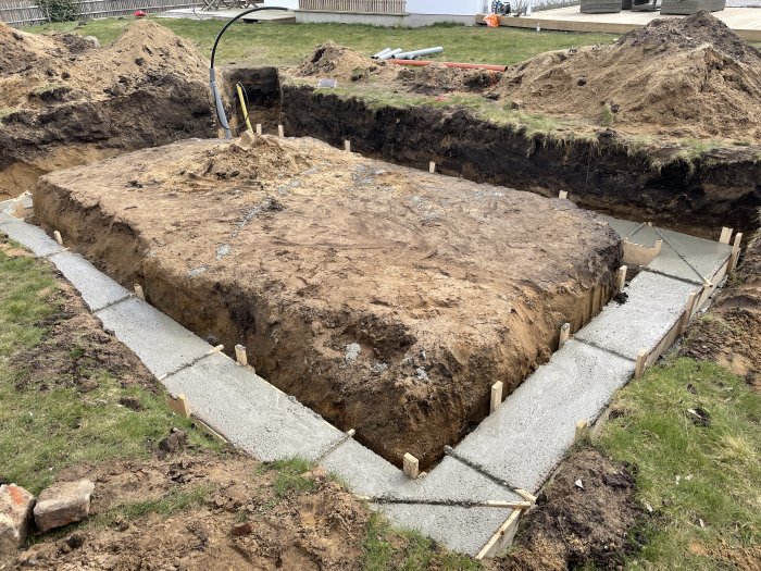 Grundarbete för växthus med gjuten sockel och utsatt jord i en trädgård.