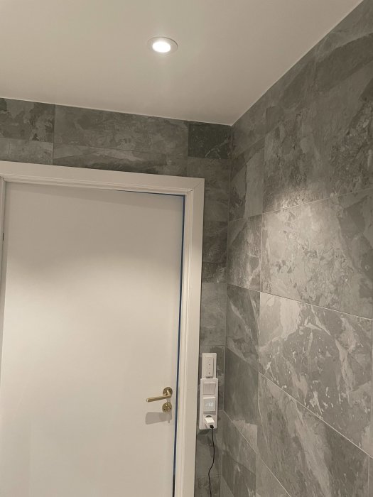 En Hide-a-lite Optic infälld takspotlight i ett badrum med grå klinker på väggarna, vit dörr och strömbrytare.