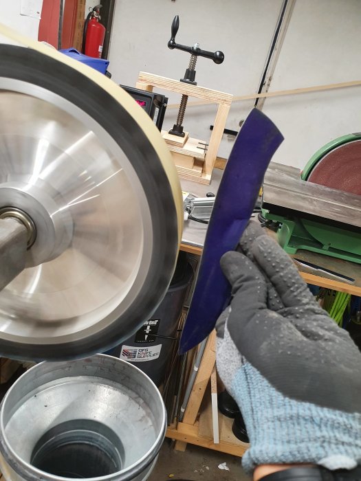 Handskeklädd hand slipar knivblad mot ett roterande kontakthjul i en verkstad.
