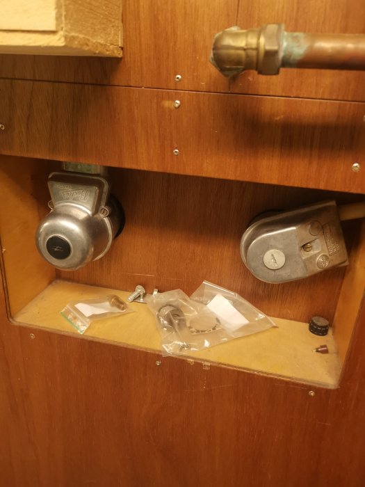 Två metalliska enheter monterade på en träpanel, en med spår för skruvmejsel och en kopplingsbox, intill kopparledningar.