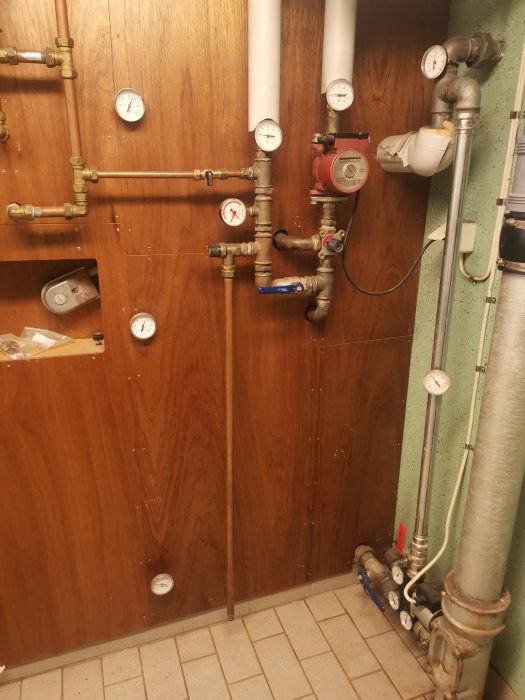 Värmesystem med kopplingsbox, termostat, rör, ventiler och manometrar på en träpanelvägg, möjlig elpatron synlig.